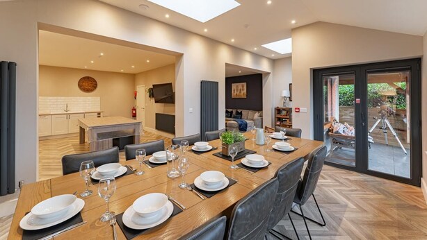Een moderne eethoek met een lange houten tafel voor zes personen, een keuken op de achtergrond en een glazen schuifdeur naar een buitenruimte.