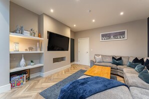 Ein modernes Wohnzimmer mit Flachbildfernseher, grauem Sofa, Couchtisch aus Holz und einem Parkettboden mit Fischgrätenmuster.