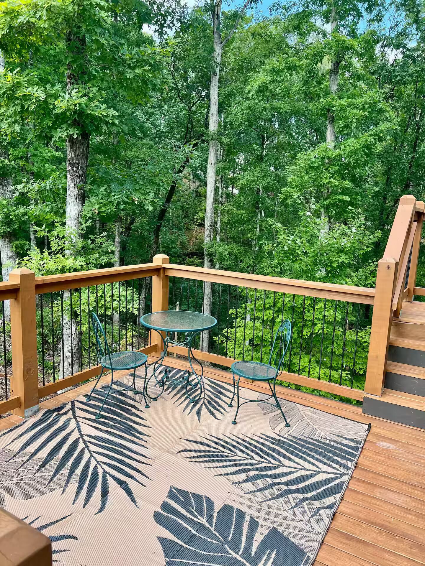 Une terrasse en bois avec une table et des chaises en verre, entourée d’une rambarde en métal, donnant sur une zone boisée.
