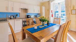 A kitchen with white cabinets, a dining table with chairs, a vase with flowers, and a view through a glass door. - 3 Bed in Ambleside (oc-lr214) (Claife)