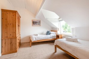 Ein Schlafzimmer mit schrägen Wänden, einem Holzschrank, zwei Betten, einem Nachttisch und einem Fenster mit Blick auf Grünflächen.