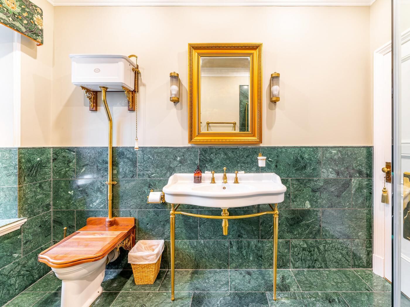 A bathroom with a marble floor, a white sink with gold fixtures, a wooden bidet, and a gold-framed mirror.