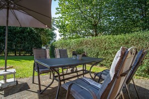 An outdoor patio with a table and chairs, a large umbrella, and a hedge in the background. - Bungalow Boddenhaus with Private Terrace, Garden, and Wi-Fi (Sundhagen)