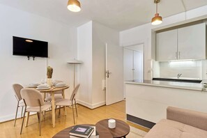 Un salon moderne comprenant un coin repas, un téléviseur à écran plat fixé au mur, une cuisine avec un plan de travail et un évier, ainsi qu’un canapé.