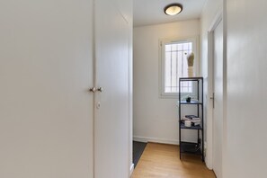 Un pasillo con piso de madera, un estante de metal negro con libros y un jarrón, y una ventana con persianas.