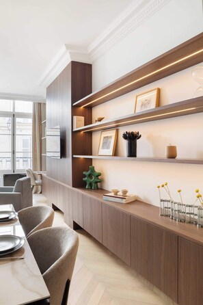 A modern hotel room with built-in wooden shelving, a dining area with chairs and a table, and a view through a window. - Luxurious Apartment 1Bdr Saint Germain des Prés (Paris)