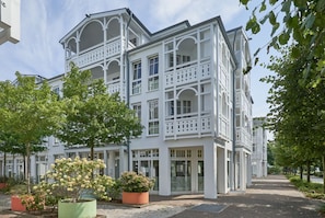 Ein weißes Gebäude mit Balkonen und großen Fenstern, umgeben von Bäumen und einem gepflasterten Weg.