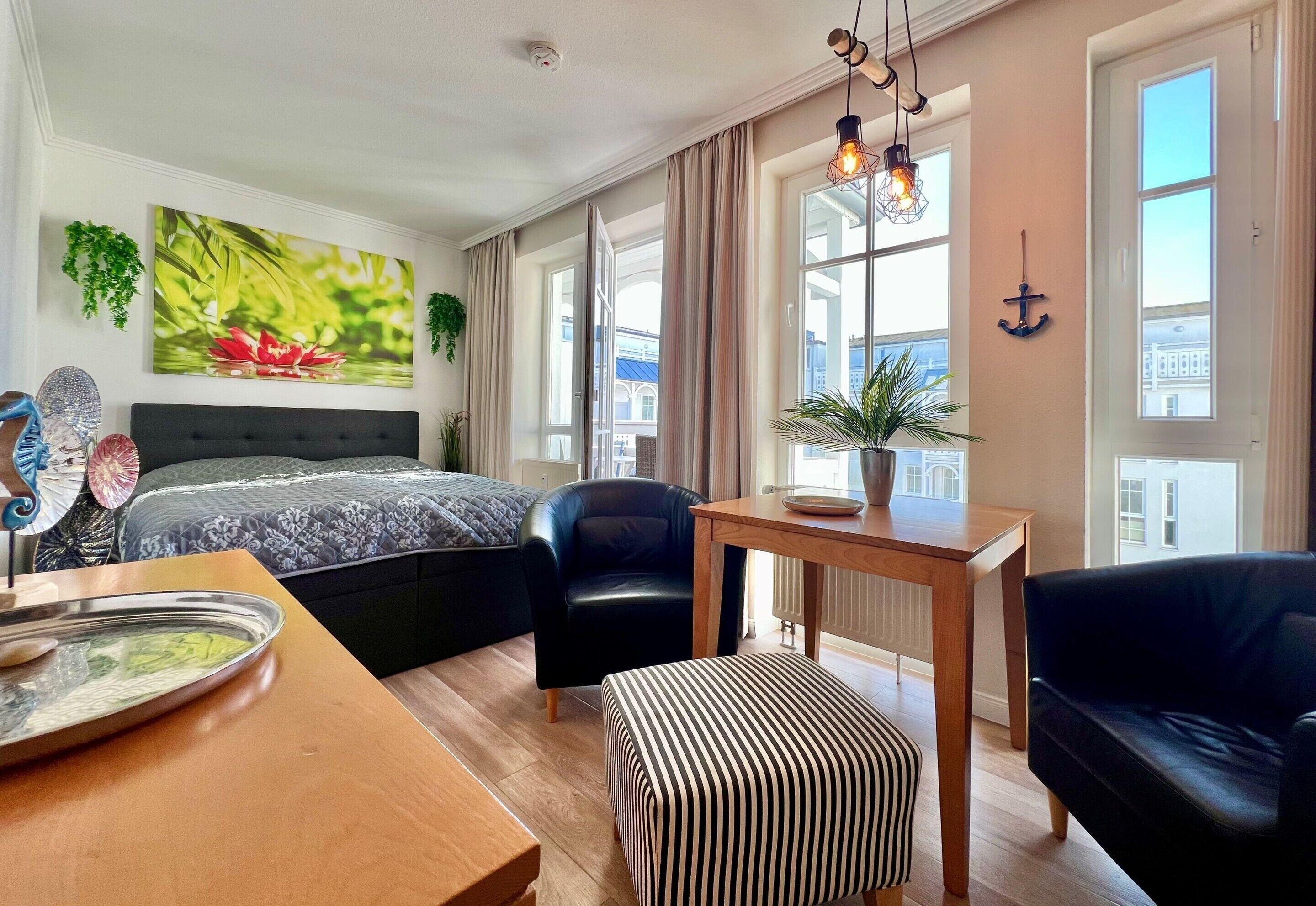 A modern hotel room with a large bed, a dining table, a black leather chair, a striped ottoman, and a view of a building through the windows.