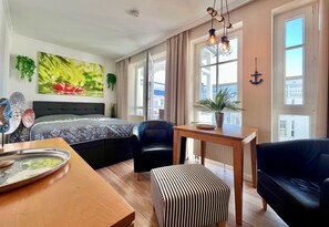 A modern hotel room with a large bed, a dining table, a black leather chair, a striped ottoman, and a view of a building through the windows. - Kompakte Ferienwohnung im Seepark Sellin mit Balkon (Sellin)