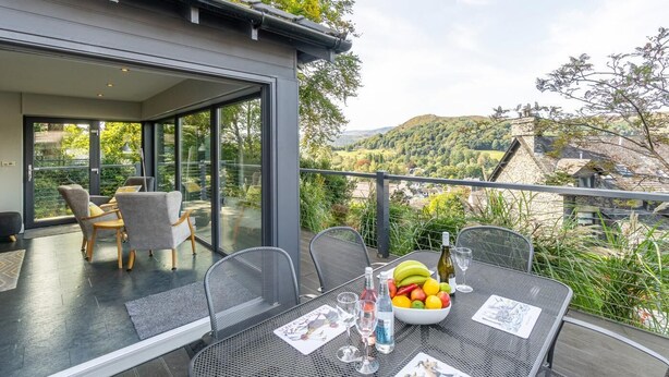 Un área de comedor exterior moderna con pared de vidrio, una mesa dispuesta con frutero, copas de vino y una carta.