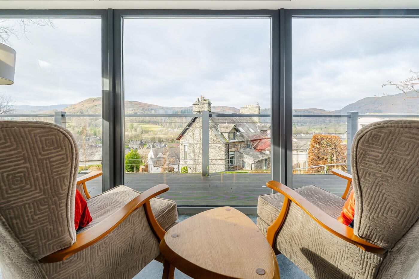 Una habitación con dos sillas y una mesita, ventanas amplias con vistas a un edificio de piedra y colinas.