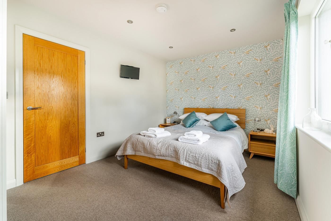 A bedroom with a wooden bed, a nightstand, a wall-mounted TV, and a patterned wallpaper.