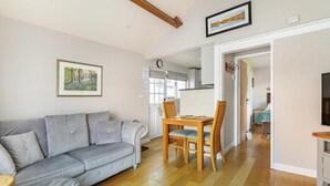 Een woonkamer met een grijze bank, een houten eettafel en stoelen, een keuken op de achtergrond en een gang die naar een andere kamer leidt.