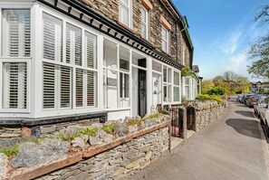 A stone building with white shutters, a stone wall, and a paved road with parked cars. - 3 Bed in Windermere (oc-lr195) (Windermere)