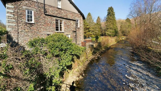 A stone building with a river in front, surrounded by trees and vegetation. - 3 Bed in Grasmere (oc-lr193) (Grasmere)