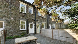 A stone building with multiple windows and doors, a gravel patio with a wooden table, and a wooden fence. - 3 Bed in Grasmere (oc-lr193) (Grasmere)