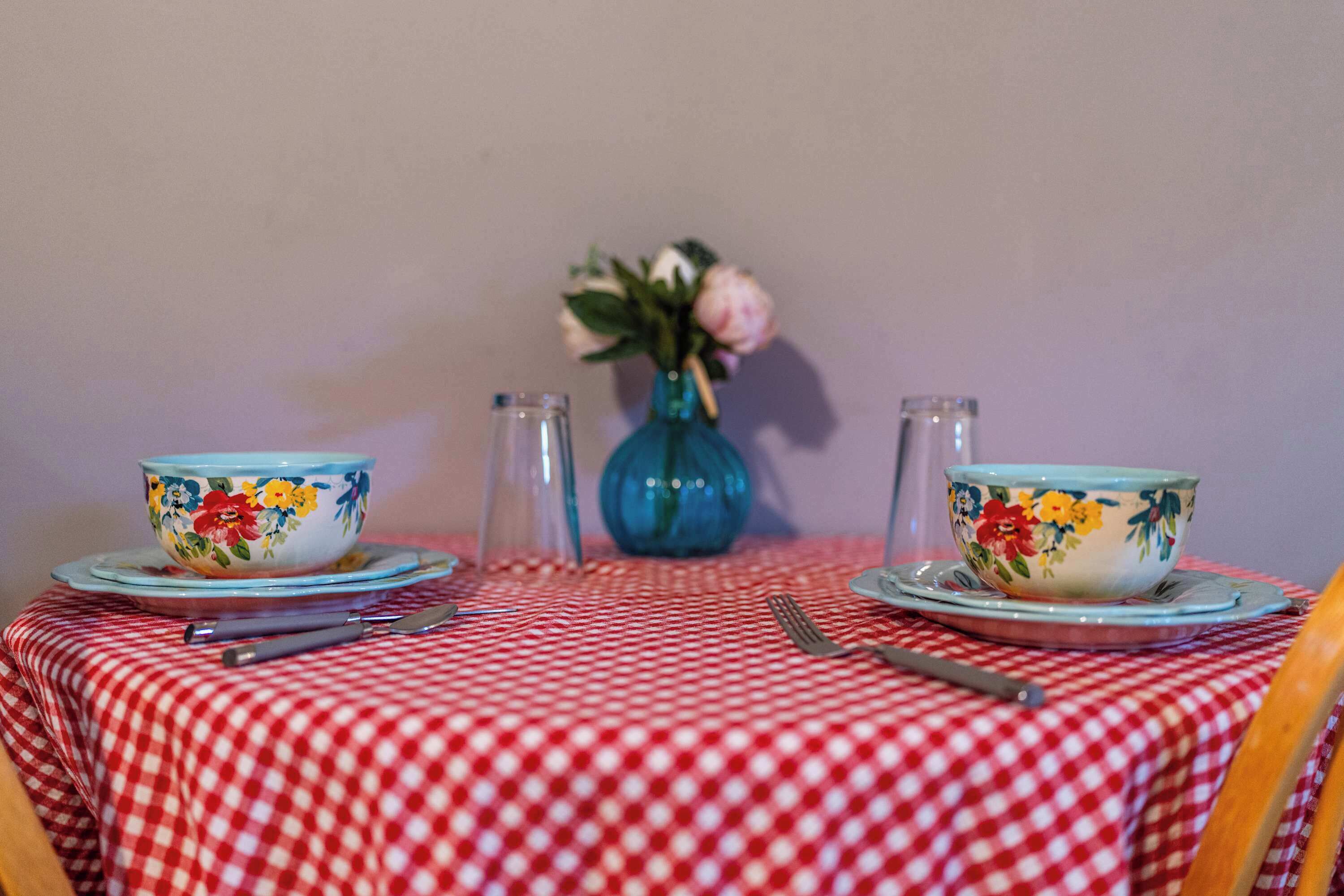 Une table à manger recouverte d’une nappe à carreaux rouges et blancs, deux bols ornés de motifs floraux, un vase avec des fleurs, des salières et des poivrières, ainsi que des couverts.