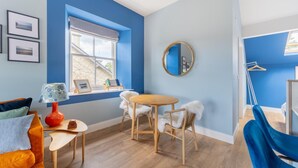 A room with a blue accent wall, a round mirror, a wooden table with chairs, a red lamp, and a window with blinds. - 1 Bed in Bowness (oc-lr150) (Bowness on Windermere)