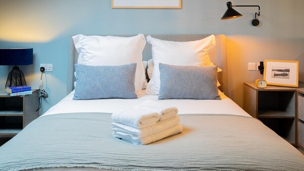 A neatly made bed with pillows, a folded towel stack, a bedside lamp, and a framed picture on a nightstand. - 1 Bed in Bowness (oc-lr150) (Bowness on Windermere)