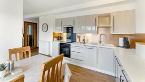 A modern kitchen with white cabinets, a built-in oven, and a sink. There is a dining area with a table and chairs. - 2 Bed in Ambleside (oc-lr191) (Ambleside)