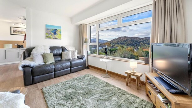 A modern living room with a leather sofa, a flat-screen TV, a large window with a scenic view, and a small side table. - 2 Bed in Ambleside (oc-lr191) (Ambleside)