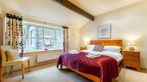 Een slaapkamer met een bed, een stoel, een nachtkastje, een raam met gordijnen en een ingelijst schilderij aan de muur.