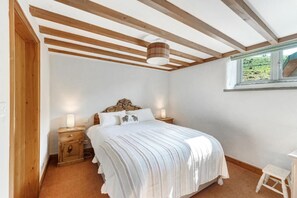 Ein Schlafzimmer mit Holzboden, einem Bett mit weißer Bettwäsche, einem Nachttisch, einer Lampe und einem Fenster mit Blick auf Grünflächen.