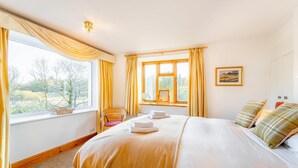 Um quarto com uma cama, duas janelas com cortinas, uma cadeira, uma mesa de cabeceira com abajur e um quadro na parede.