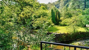Ein üppiger Garten mit einer Steinbrücke, dichtem Laub und freiem Blick auf eine Hügelkulisse im Hintergrund.