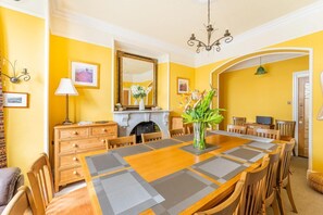 A dining room with a long wooden table, chairs, a vase with flowers, a fireplace, and a mirror on the wall. - 7 Bed in Ambleside (oc-lr053) (Ambleside)