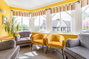 A room with a yellow wall, a grey sofa, two yellow armchairs, a potted plant, and large windows with curtains. - 7 Bed in Ambleside (oc-lr053) (Ambleside)