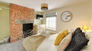 Une chambre avec une cheminée en briques, un lit avec de nombreux coussins, un miroir rond et une lampe de chevet.