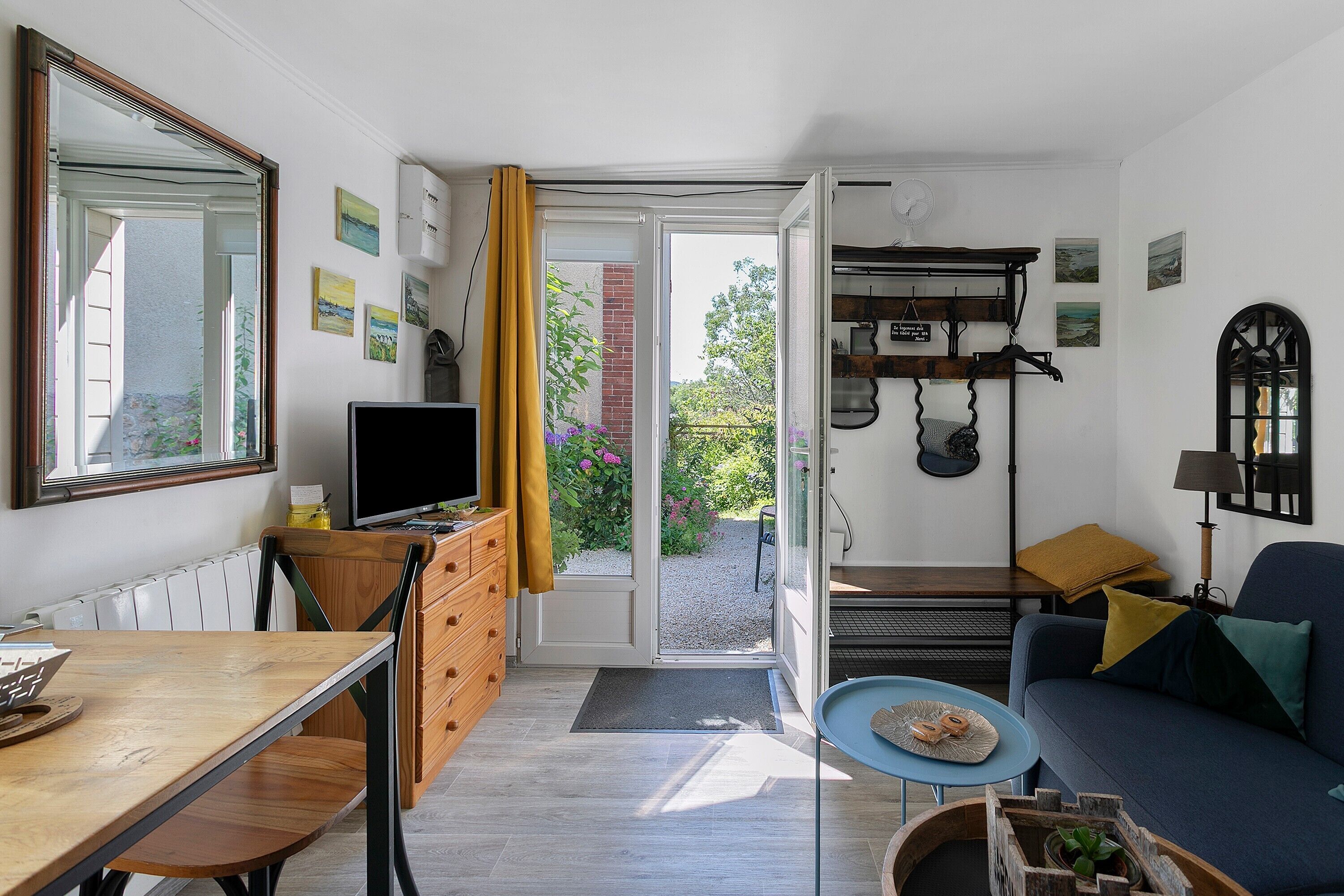Un espace de vie compact comprenant un coin repas, un canapé, un téléviseur et une vue sur un jardin, accessible par une porte ouverte.