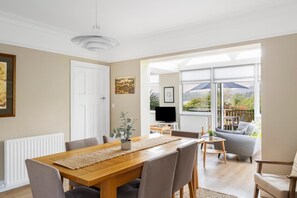 A dining area with a wooden table and chairs, a television, and a view of a garden through large windows. - 4 Bed in Ambleside (oc-lr084) (Ambleside)