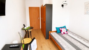 Ein kleiner, gut beleuchteter Raum mit einem Bett, einem Schreibtisch mit Stuhl, einem Tisch mit Stuhl, einem Fernseher und einer Tür.