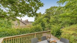 Eine Holzterrasse mit Tisch und Stühlen, umgeben von Grünpflanzen und einem Steinhaus im Hintergrund.