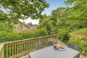 A wooden deck with a table and chairs, surrounded by greenery and a stone building in the background. - 3 Bed in Hawkshead (oc-lr046) (Claife)
