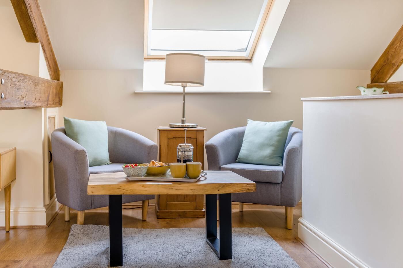 Une pièce chaleureuse avec une lucarne, deux fauteuils gris, une table en bois sur laquelle se trouve un plateau de nourriture et une lampe sur pied.