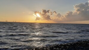 En kustvy med havet, en stenig strand och avlägsna industriella byggnader.