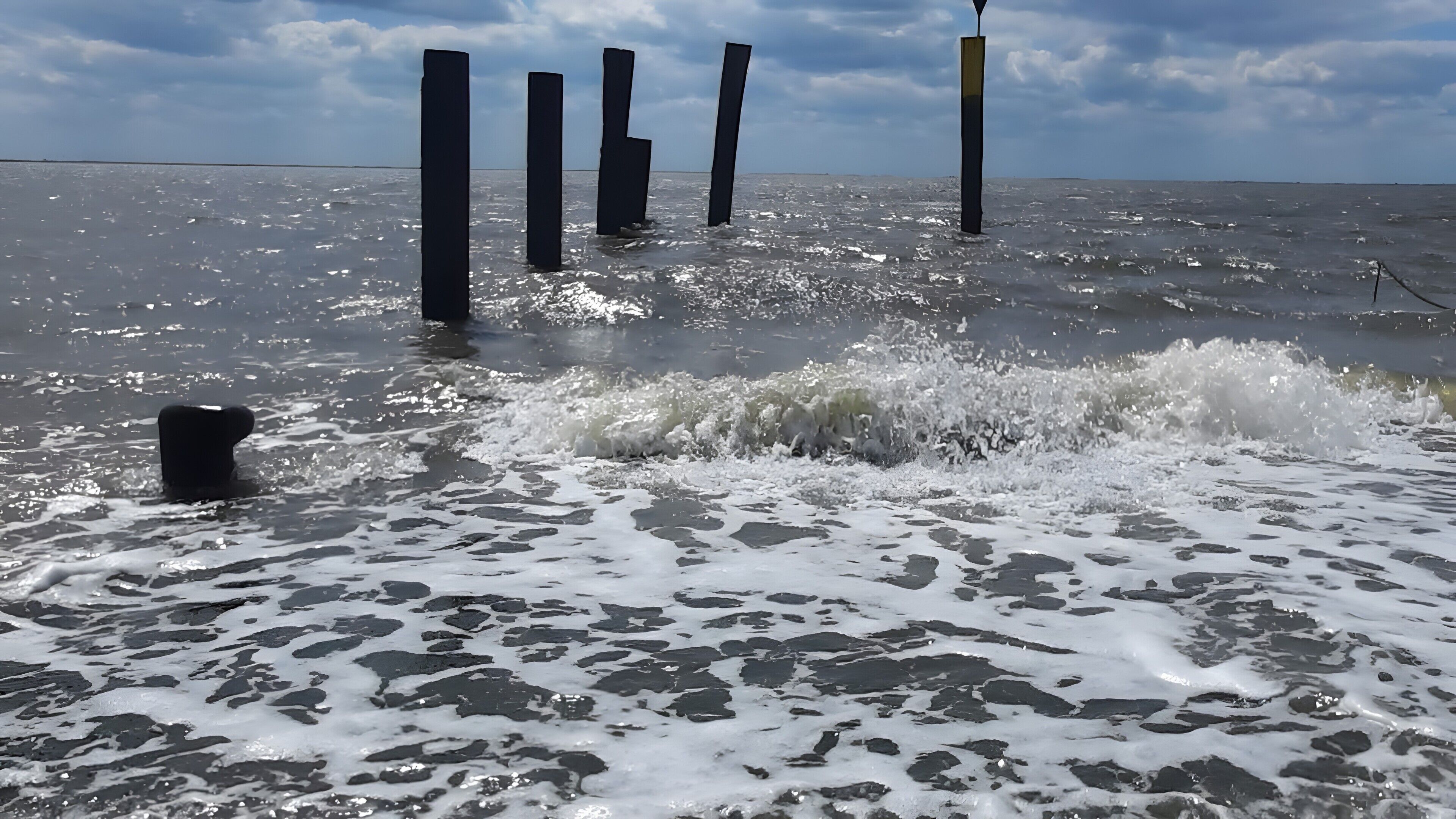 Ein Strand mit brechenden Wellen und mehreren senkrechten Pfählen im Wasser.