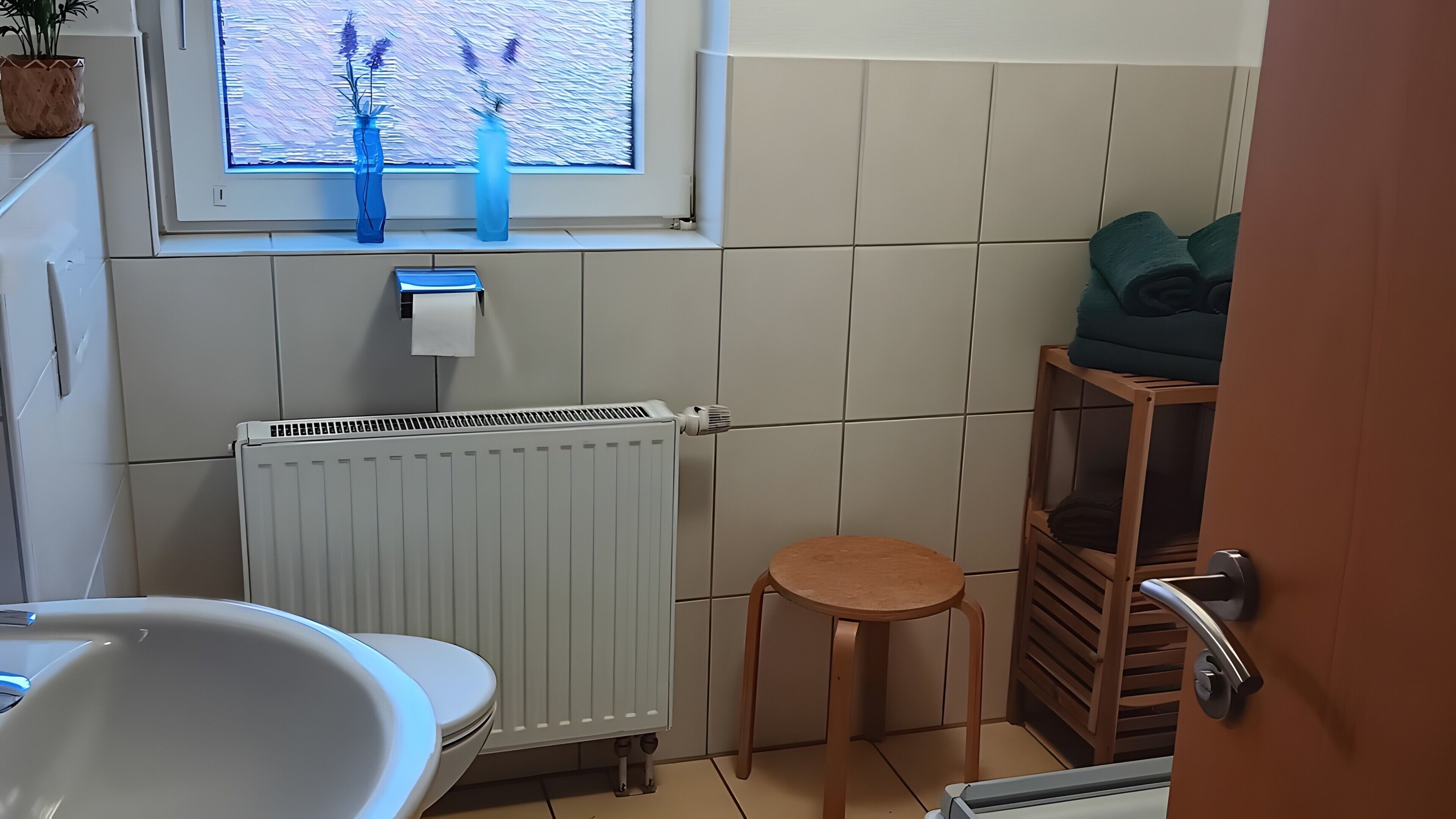 Un baño con un radiador blanco, una ventana con vistas a plantas de lavanda, un inodoro y un pequeño taburete de madera.