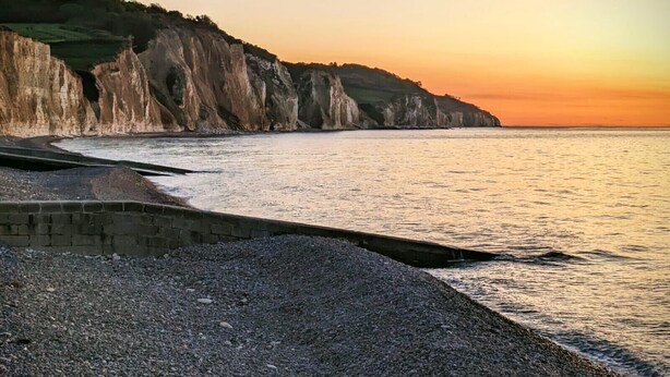 Un paisaje costero con acantilados, una playa de guijarros y un mar tranquilo al atardecer.
