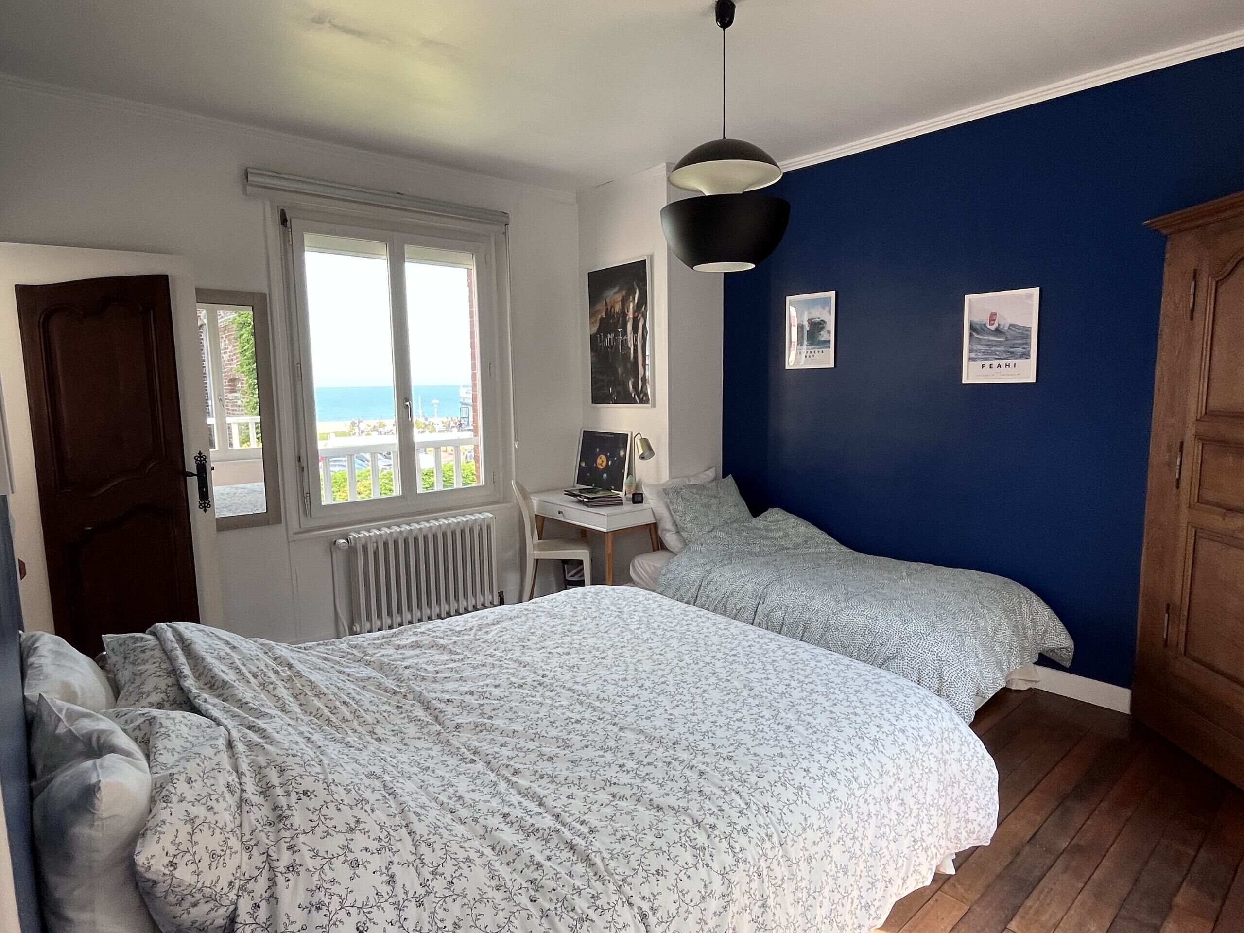 A bedroom with two beds, a desk, a radiator, a window with a view, and a door.
