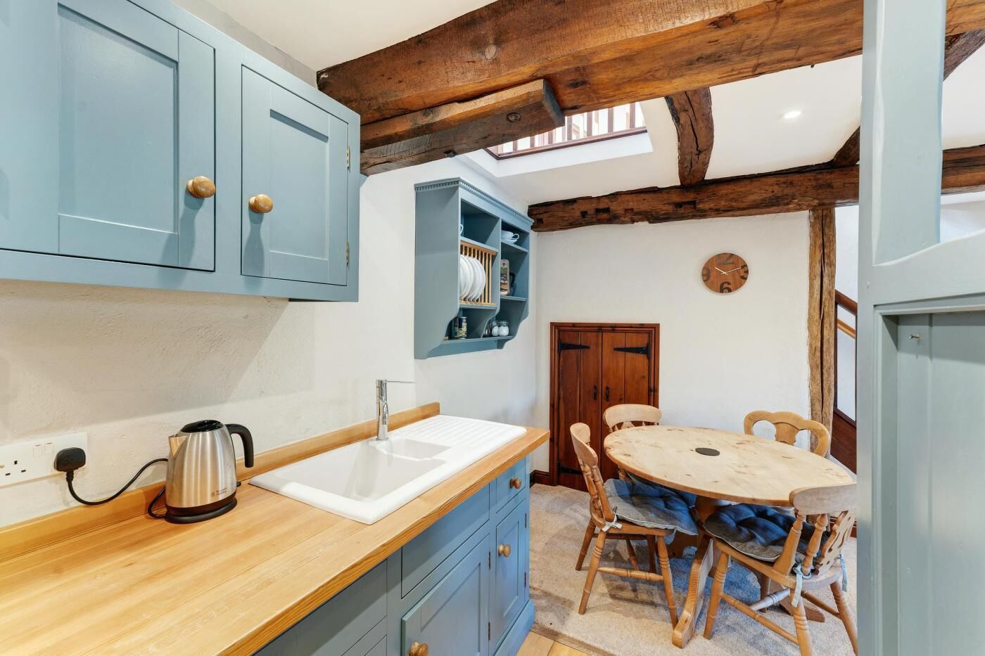 Une cuisine avec un plan de travail en bois, un évier et une bouilloire. On y trouve une table à manger avec des chaises et une horloge au mur.