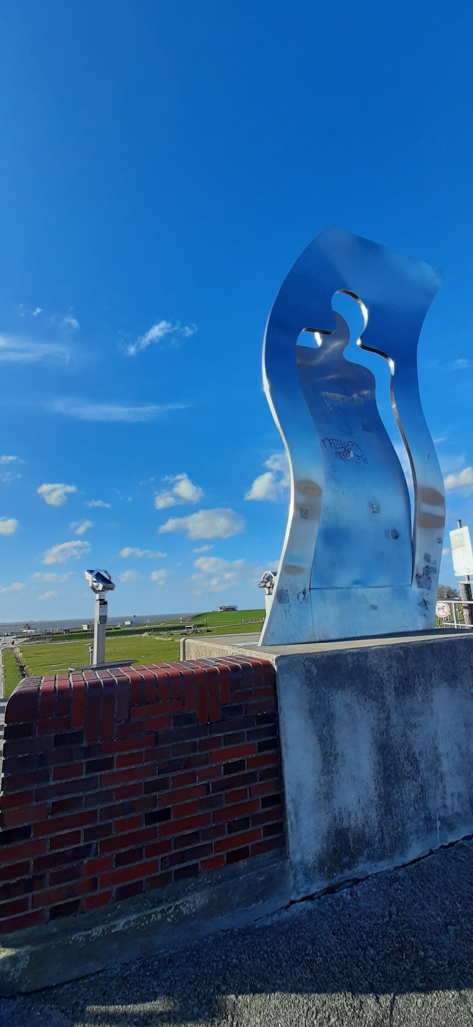 Een metalen sculptuur met een uitgesneden vorm, een straatlamp op zonne-energie, en een bakstenen muur.