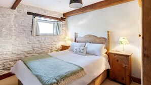 Een slaapkamer met een stenen muur, houten balken, een bed met witte beddengoed, twee nachtkastjes met lampen en een raam met gordijnen.