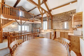 Un interior rústico con una mesa de comedor de madera, una cocina con paredes de piedra y un dormitorio en entrepiso con escalera de madera.