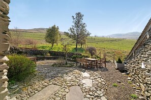 A stone patio with a table and chairs, surrounded by greenery and a stone wall. - 2 Bed in Ambleside (oc-lr177) (Ambleside)
