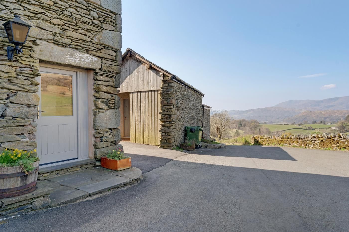 A stone building with a white door, a wooden door, and a paved driveway leading to a rural landscape.
