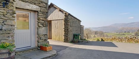 A stone building with a white door, a wooden door, and a paved driveway leading to a rural landscape.